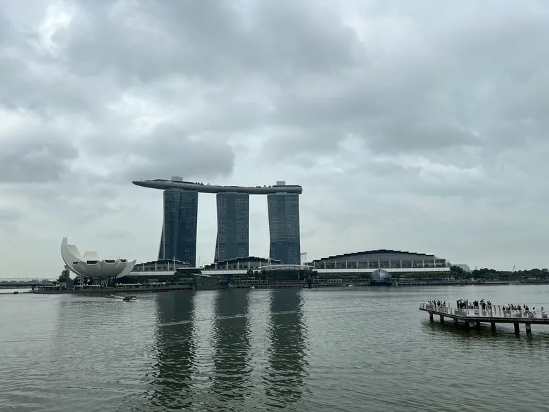 Marina Bay in Singapore with the three-tower structure of Marina Bay Sands topped by its long rooftop deck, the lotus-shaped ArtScience Museum to the left, and the waterfront promenade to the right where groups of people stand on an extended viewing platform above the water. Reflections ripple across the calm bay under a cloudy, gray sky.