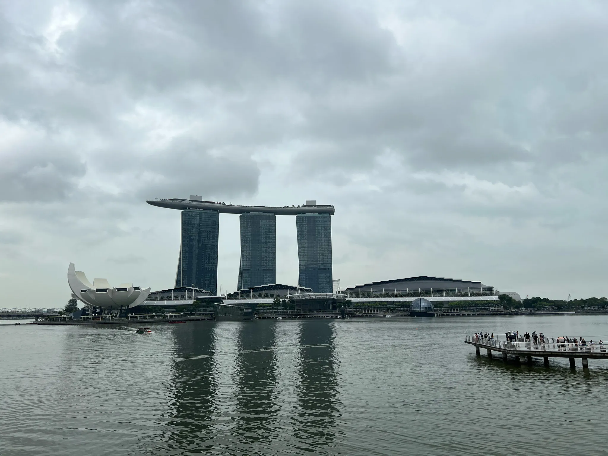 Marina Bay in Singapore with the three-tower structure of Marina Bay Sands topped by its long rooftop deck, the lotus-shaped ArtScience Museum to the left, and the waterfront promenade to the right where groups of people stand on an extended viewing platform above the water. Reflections ripple across the calm bay under a cloudy, gray sky.