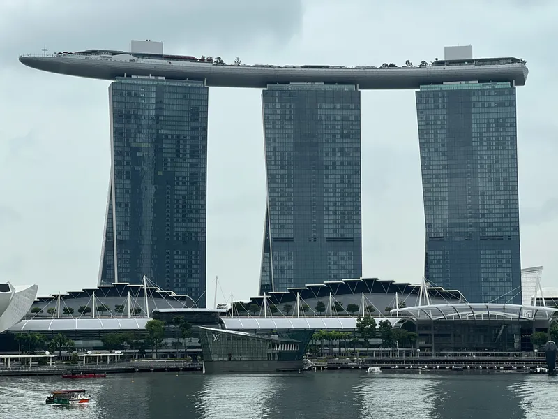 Three tall, modern high‑rise towers rise side by side, each clad in reflective blue‑gray glass. A long, curved rooftop structure resembling a ship spans across the tops of the towers, forming a dramatic elevated platform. Below the towers lies a complex of low, angular buildings with white structural supports and glass facades. In the foreground, calm water reflects the skyline, and a small boat moves across the bay. The sky is overcast, giving the scene a soft and muted tone.