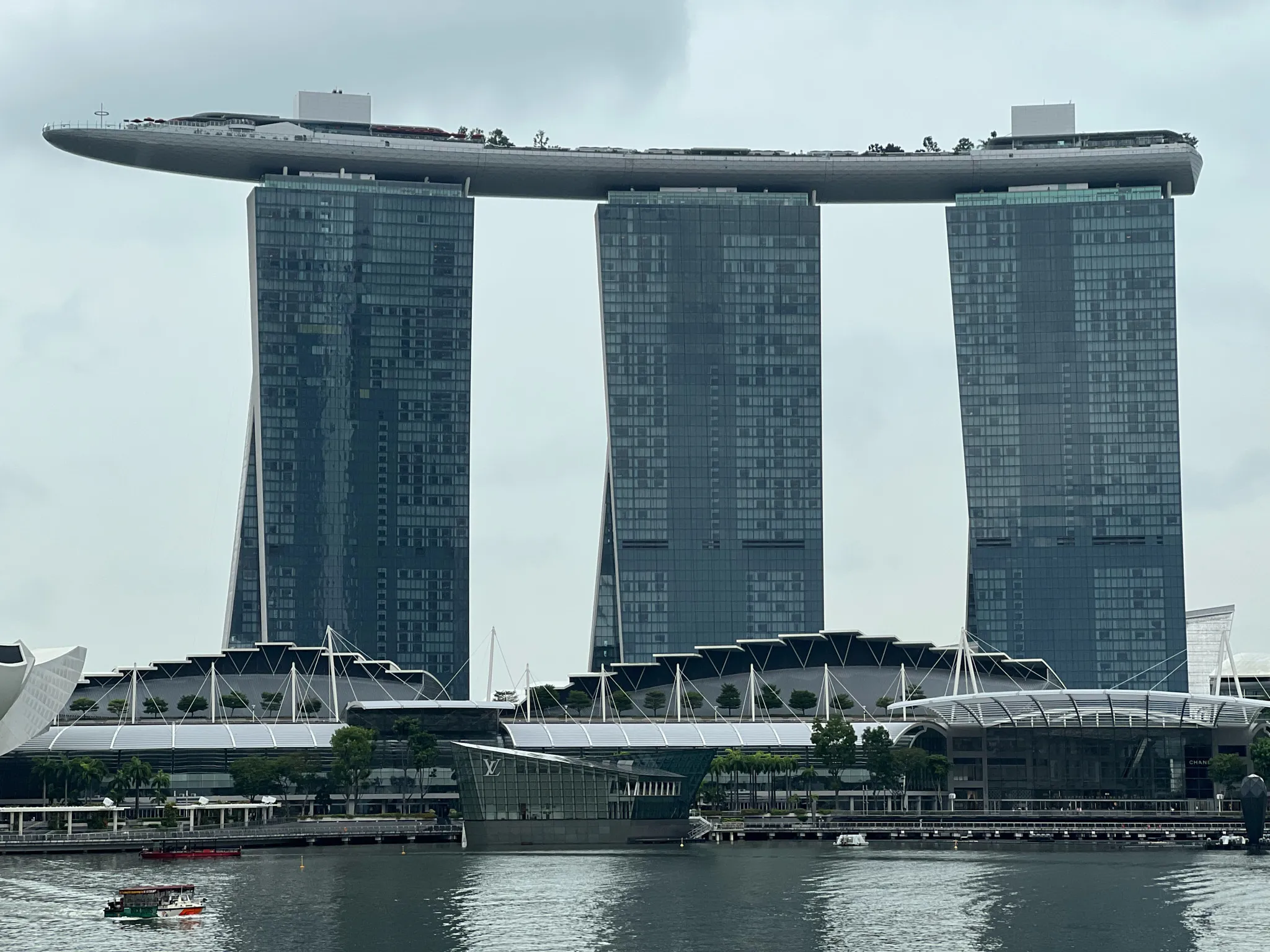 Three tall, modern high‑rise towers rise side by side, each clad in reflective blue‑gray glass. A long, curved rooftop structure resembling a ship spans across the tops of the towers, forming a dramatic elevated platform. Below the towers lies a complex of low, angular buildings with white structural supports and glass facades. In the foreground, calm water reflects the skyline, and a small boat moves across the bay. The sky is overcast, giving the scene a soft and muted tone.
