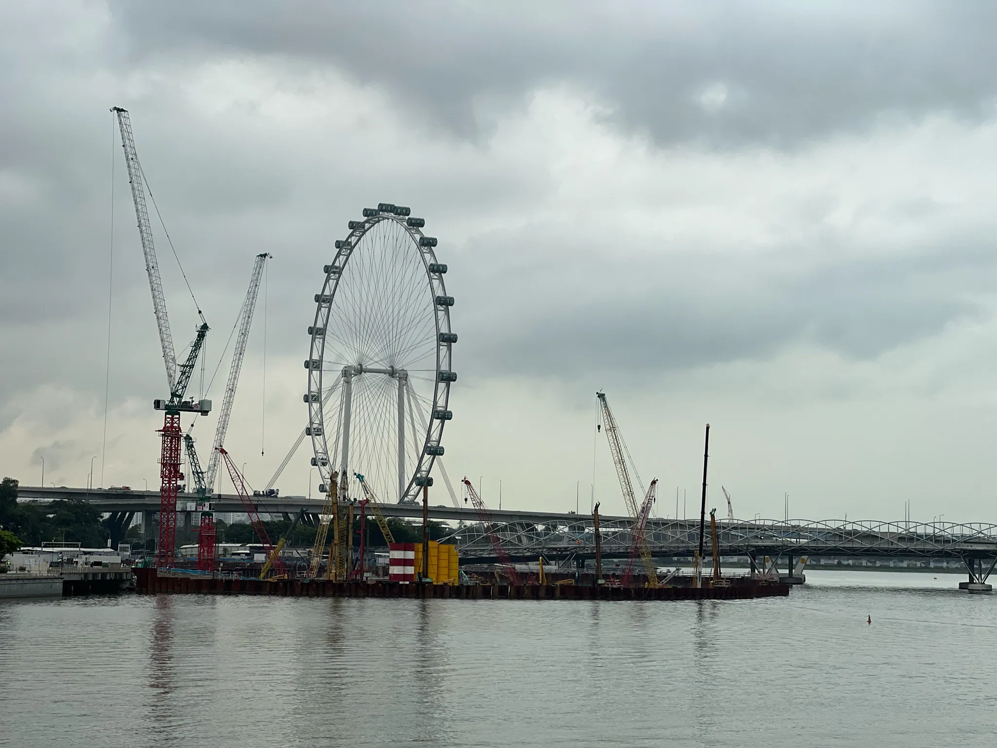 Large cranes and construction equipment line a floating work platform on calm waterfront water, set against a towering observation wheel and an overcast sky. A bridge spans the background as multiple cranes rise around the wheel, suggesting ongoing urban development along the shoreline.