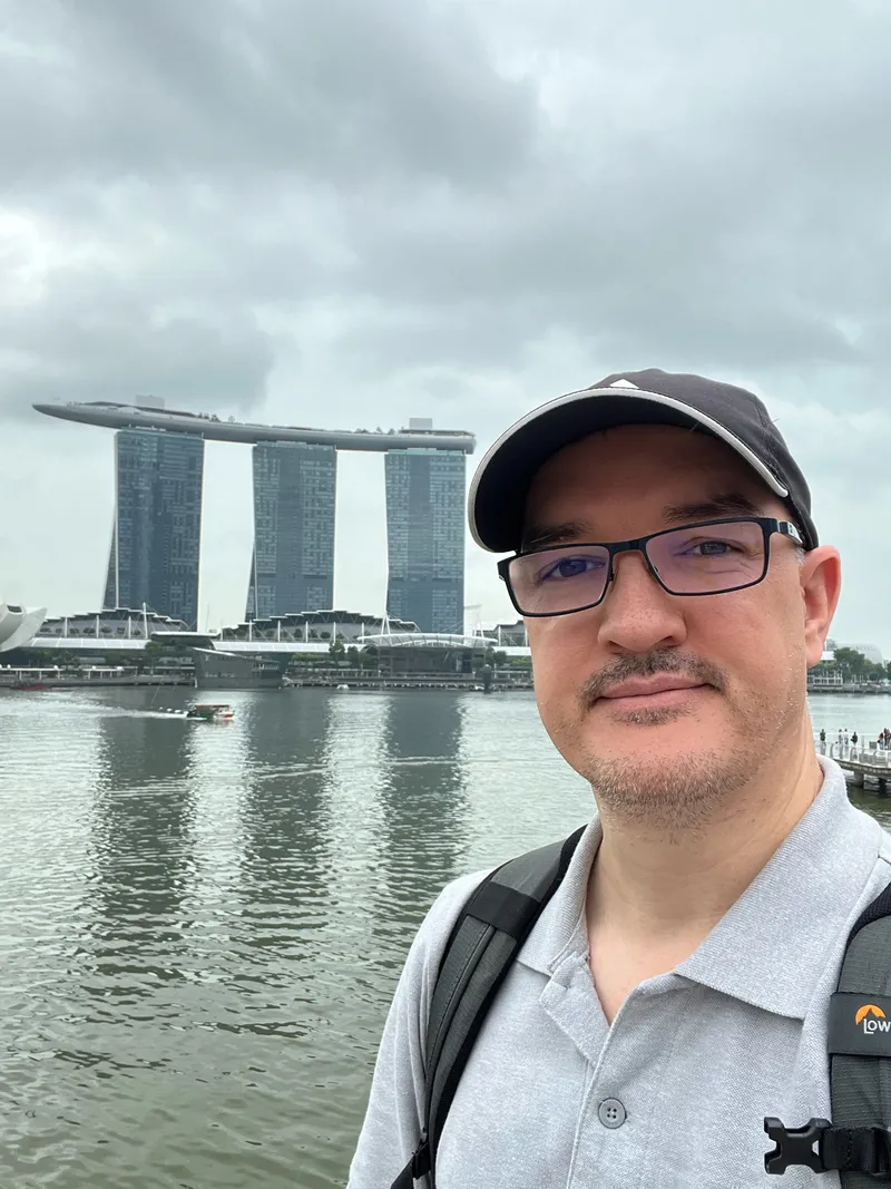 A person wearing glasses, a dark cap, and a gray polo shirt stands near a waterfront with calm, reflective water behind them. In the background rises the distinct three‑tower structure topped by a long platform, part of a modern urban skyline. The sky is overcast, creating soft, diffused light over the water and surrounding buildings.