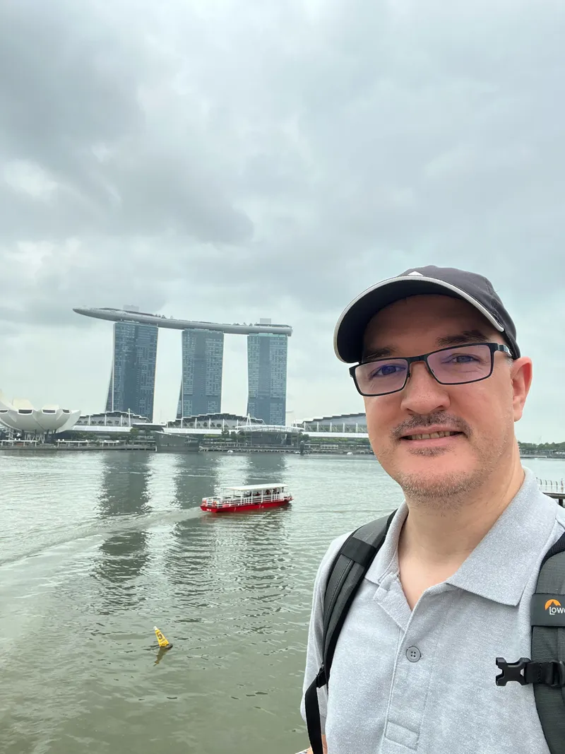 A person wearing glasses, a dark cap, and a grey polo shirt stands near a waterfront with calm water in the foreground. A small red boat moves across the bay, leaving a gentle wake behind. Across the water, a distinctive three‑tower building topped by a long horizontal structure rises beneath an overcast sky, with additional modern buildings and a dome-shaped structure nearby.
