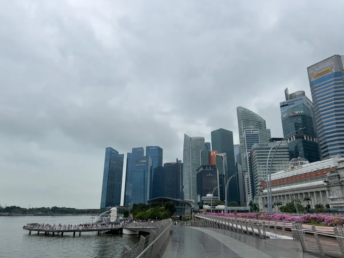 In the Civic District, Downtown Core, Central Region of Singapore, an impressive skyline of futuristic skyscrapers stretches beneath a cloudy sky. At the centre of the image, a picturesque walkway gathers a crowd of visitors. At its end stands the famous Merlion, one of Singapore's iconic landmarks. Green plants in the surroundings form a pleasing contrast to the modern architecture. Clouds in the sky hint at a change in weather to come.