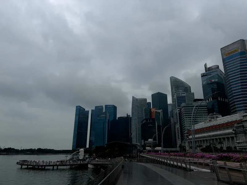 A waterfront promenade leads toward a cluster of tall, modern skyscrapers under a dark, overcast sky. Dense clouds hang low, casting a muted gray light across the scene. To the left, a viewing platform extends over the water, where a white fountain sculpture sprays upward. The skyline is filled with glass and steel towers of varying shapes, some with curved facades and others with sharp, angular lines. On the right side, a row of magenta flowers decorates the edge of the walkway beside an older building with a red roof, contrasting with the contemporary architecture behind it.
