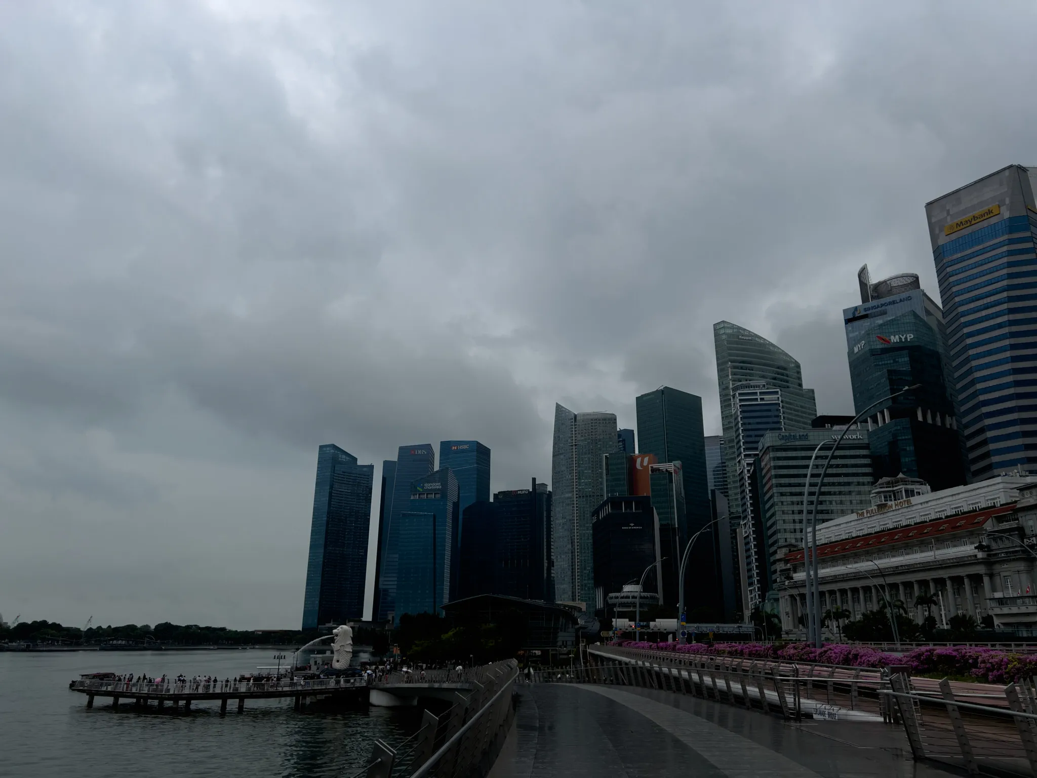 A waterfront promenade leads toward a cluster of tall, modern skyscrapers under a dark, overcast sky. Dense clouds hang low, casting a muted gray light across the scene. To the left, a viewing platform extends over the water, where a white fountain sculpture sprays upward. The skyline is filled with glass and steel towers of varying shapes, some with curved facades and others with sharp, angular lines. On the right side, a row of magenta flowers decorates the edge of the walkway beside an older building with a red roof, contrasting with the contemporary architecture behind it.