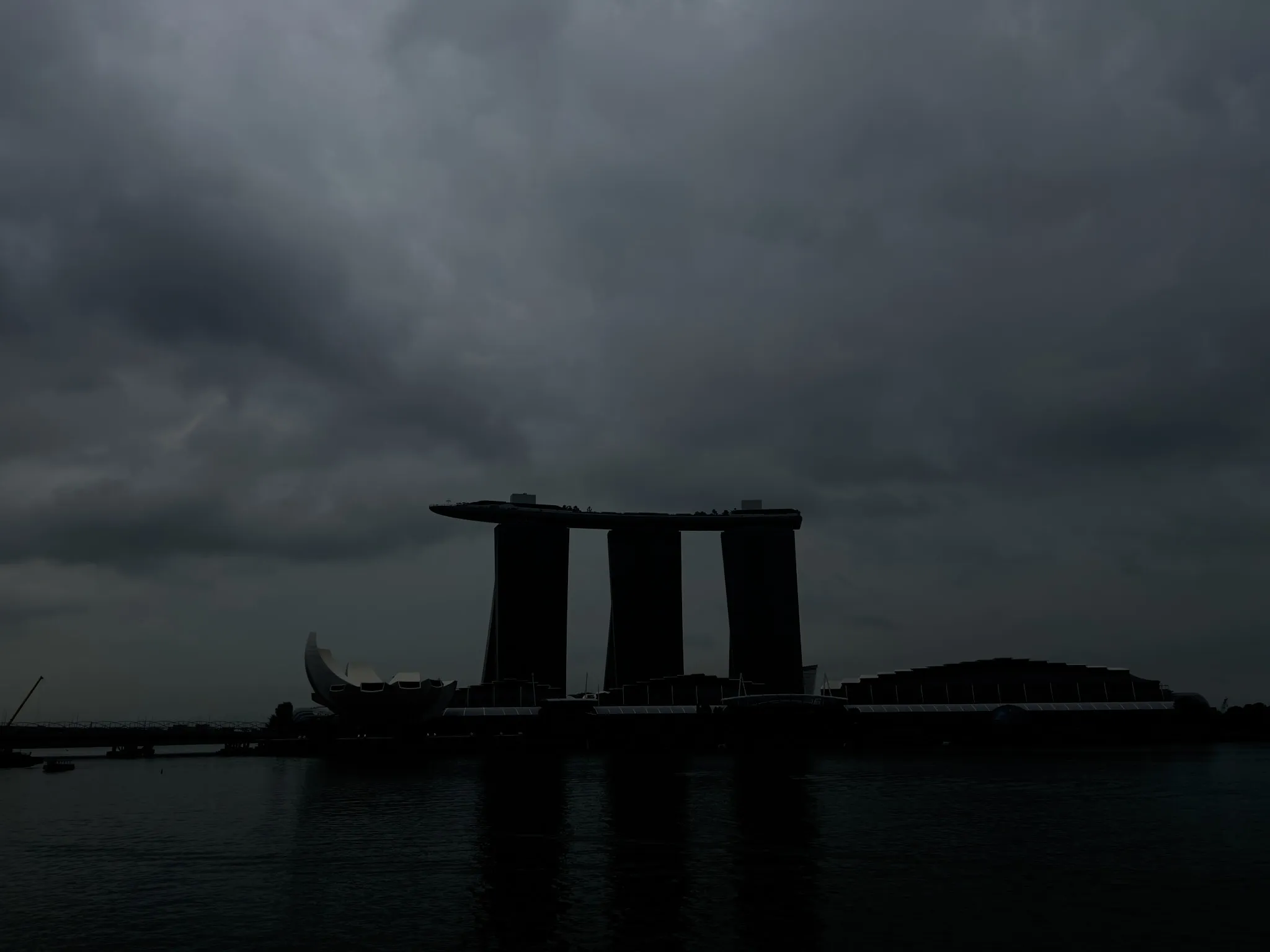 Silhouetted waterfront skyline with three tall, connected towers and a curved, lotus‑shaped building in front of them, set against a dark, overcast sky with thick storm clouds reflecting dimly on the calm water.