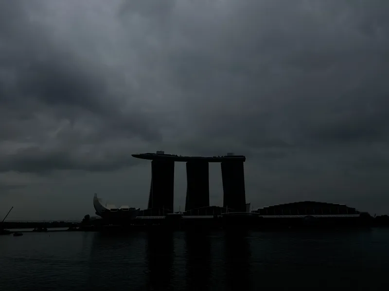 Silhouetted waterfront skyline with three tall, connected towers and a curved, lotus‑shaped building in front of them, set against a dark, overcast sky with thick storm clouds reflecting dimly on the calm water.
