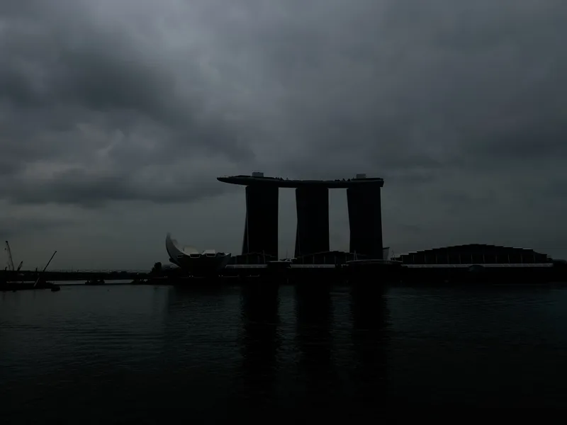 A dramatic waterfront cityscape with dark storm clouds hanging low over three tall, modern towers topped by a long, flat sky park. To the left sits a lotus-shaped building, while the structures stretch across the shoreline in deep silhouette. The still water in the foreground reflects the muted shapes beneath the overcast sky.