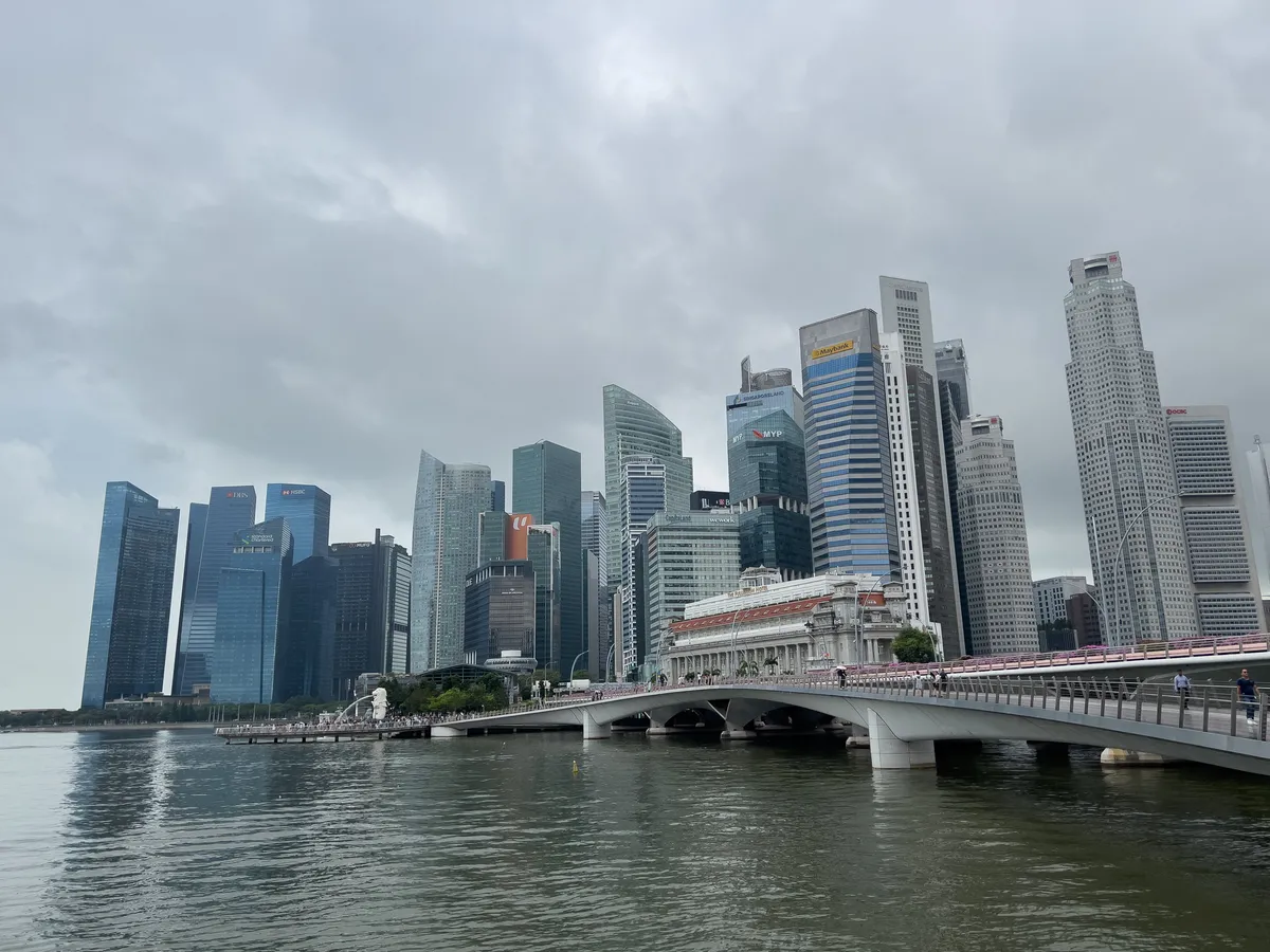 The scene shows the impressive skyline of Singapore, captured in Little India. High-rises, some glass-clad and modern, reach into the sky and reflect the light of clouds drifting above the water. At the centre, an elegant bridge spans the calm water while people cross it. In the foreground, the water surface softly mirrors the buildings and the clouds.