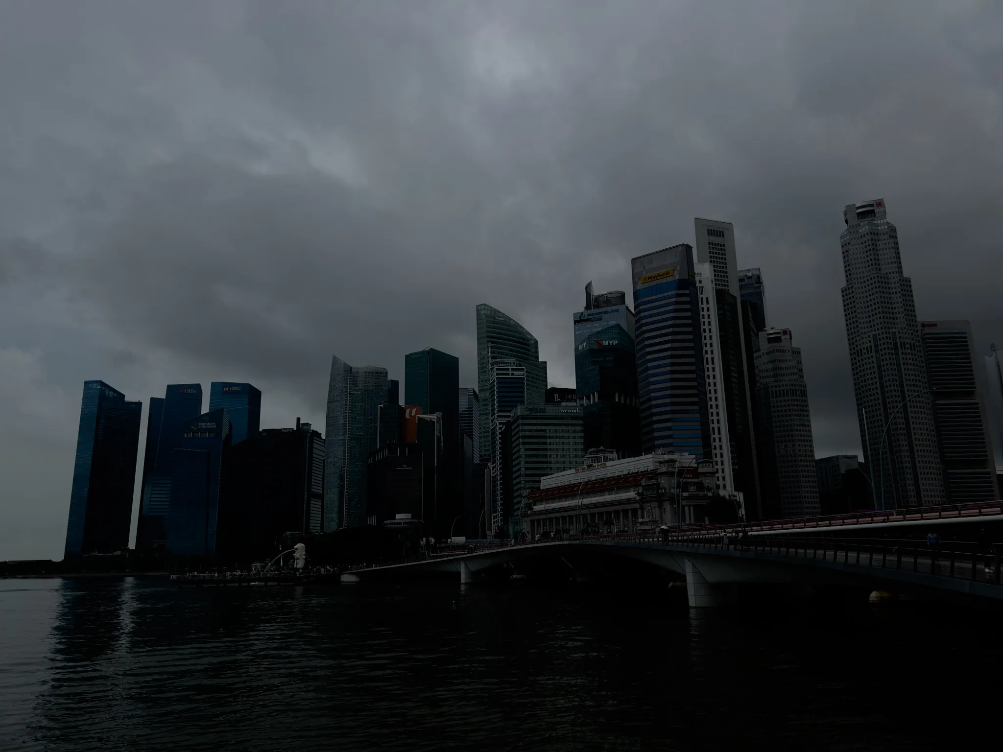 A dense cluster of modern skyscrapers rises along a waterfront under heavy, dark storm clouds. The tall buildings vary in height and design, with sleek glass facades and sharp geometric lines. A long bridge extends from the right side of the scene toward the city, running above calm, reflective water. The overall atmosphere is dim and moody, emphasizing the dramatic contrast between the towering structures and the overcast sky.