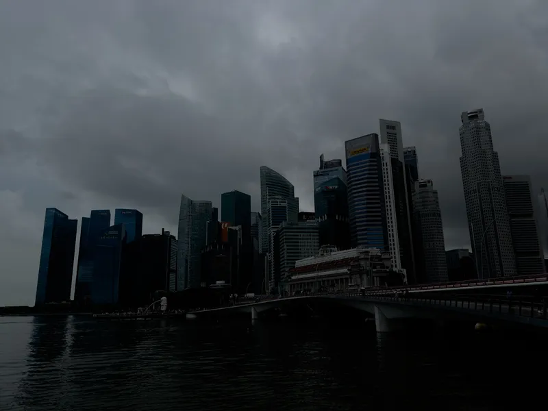 A dense cluster of modern skyscrapers rises along a waterfront under heavy, dark storm clouds. The tall buildings vary in height and design, with sleek glass facades and sharp geometric lines. A long bridge extends from the right side of the scene toward the city, running above calm, reflective water. The overall atmosphere is dim and moody, emphasizing the dramatic contrast between the towering structures and the overcast sky.