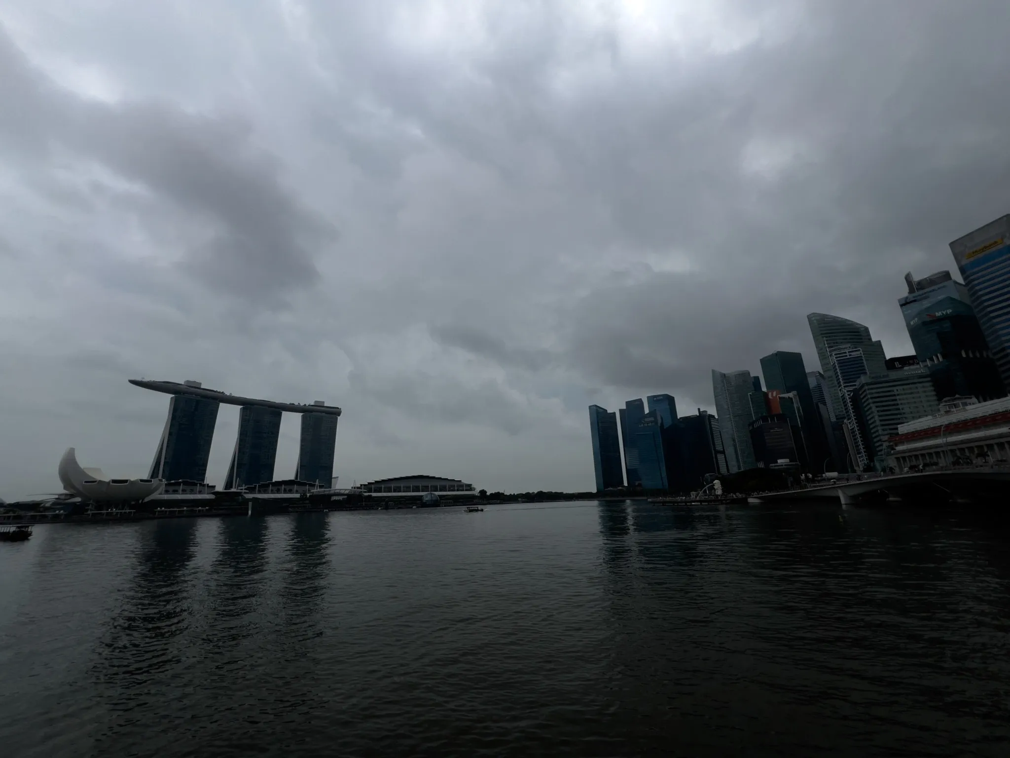 A waterfront cityscape under a heavily overcast sky, with dark clouds stretching across the scene. On the left, a distinctive three-tower structure topped by a long, curved platform rises beside a lotus-shaped building near the water. To the right, a dense cluster of tall, modern skyscrapers lines the shore. The calm bay reflects the muted tones of the buildings and sky, creating a moody, atmospheric urban view.
