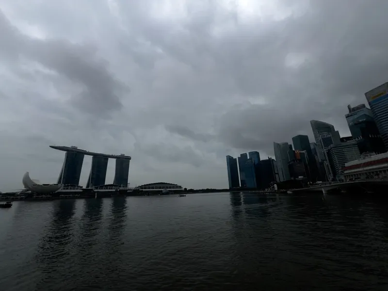 A waterfront cityscape under a heavily overcast sky, with dark clouds stretching across the scene. On the left, a distinctive three-tower structure topped by a long, curved platform rises beside a lotus-shaped building near the water. To the right, a dense cluster of tall, modern skyscrapers lines the shore. The calm bay reflects the muted tones of the buildings and sky, creating a moody, atmospheric urban view.