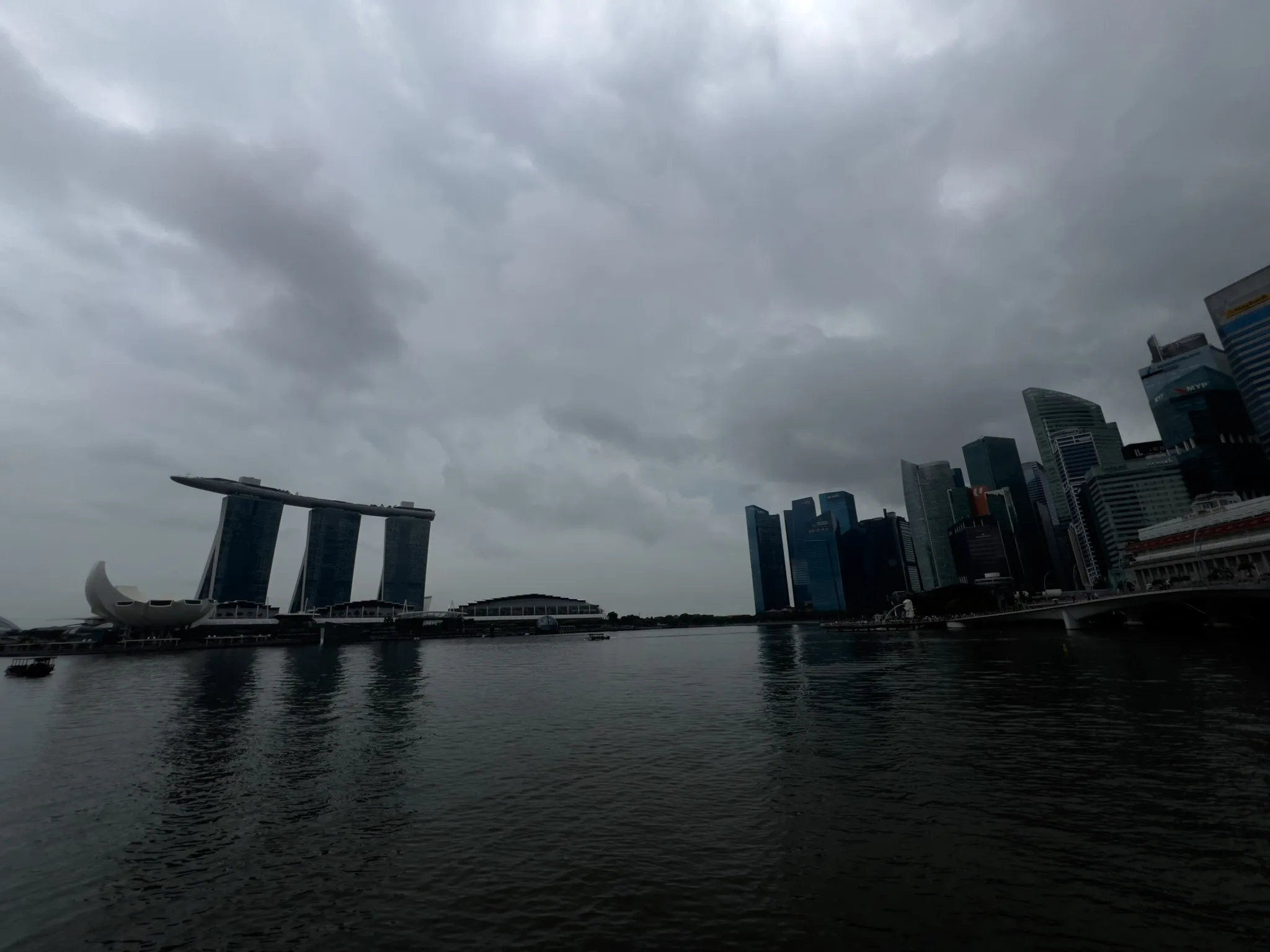 A waterfront cityscape under a dark, heavily overcast sky, with calm water in the foreground reflecting the gray clouds. On the left stands a distinctive building complex with three tall towers supporting a horizontal structure, accompanied by a rounded, lotus-like building near the shore. On the right, a cluster of modern high‑rise buildings forms a dense urban skyline. The overall atmosphere is moody and dim due to the thick cloud cover.