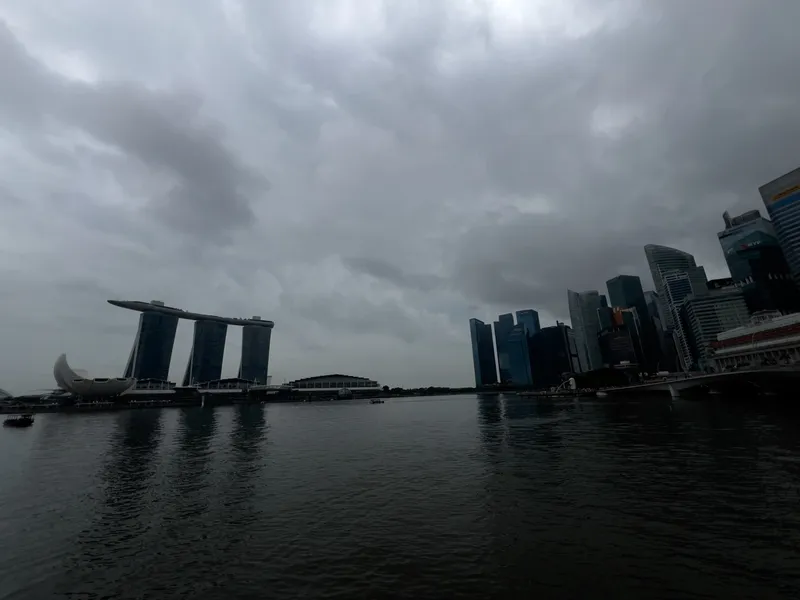 A waterfront cityscape under a dark, heavily overcast sky, with calm water in the foreground reflecting the gray clouds. On the left stands a distinctive building complex with three tall towers supporting a horizontal structure, accompanied by a rounded, lotus-like building near the shore. On the right, a cluster of modern high‑rise buildings forms a dense urban skyline. The overall atmosphere is moody and dim due to the thick cloud cover.