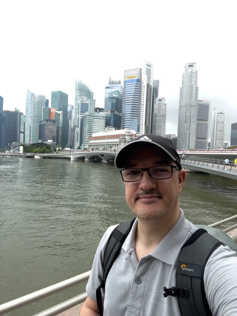 A man wearing glasses, a dark cap, and a grey polo shirt stands near a railing beside a large body of water, with modern high‑rise buildings forming a dense city skyline in the background. A bridge extends across the water behind him, and the sky is overcast, creating soft, diffused lighting over the urban scene.