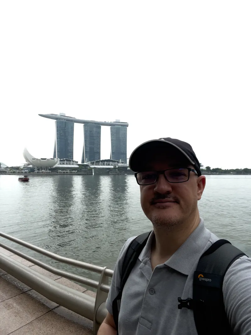 A man wearing glasses, a cap, and a backpack stands near a waterfront railing with calm water behind him. Across the water rises a distinctive three‑tower structure topped by a long, curved platform, with a lotus‑shaped building nearby. The sky is overcast, and a small boat is visible on the water to the left.