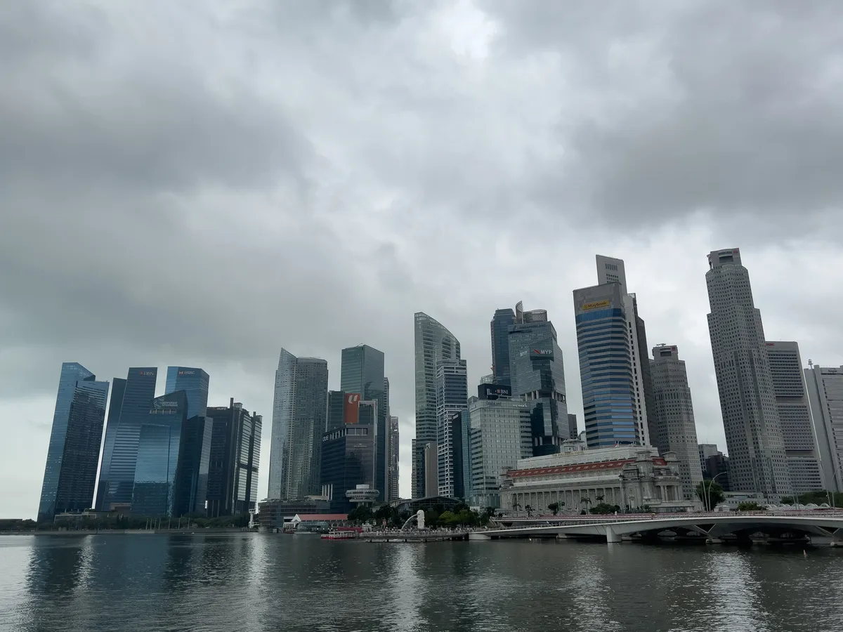An impressive Singapore skyline, captured in Little India, shows a variety of modern high-rises stretching along the water. The buildings are mirrored in the calm surface while the sky is covered by dense grey clouds hinting at imminent rain. A historic structure in the foreground sets itself apart from the modern skyscrapers, forming a stark contrast with the contemporary architecture.