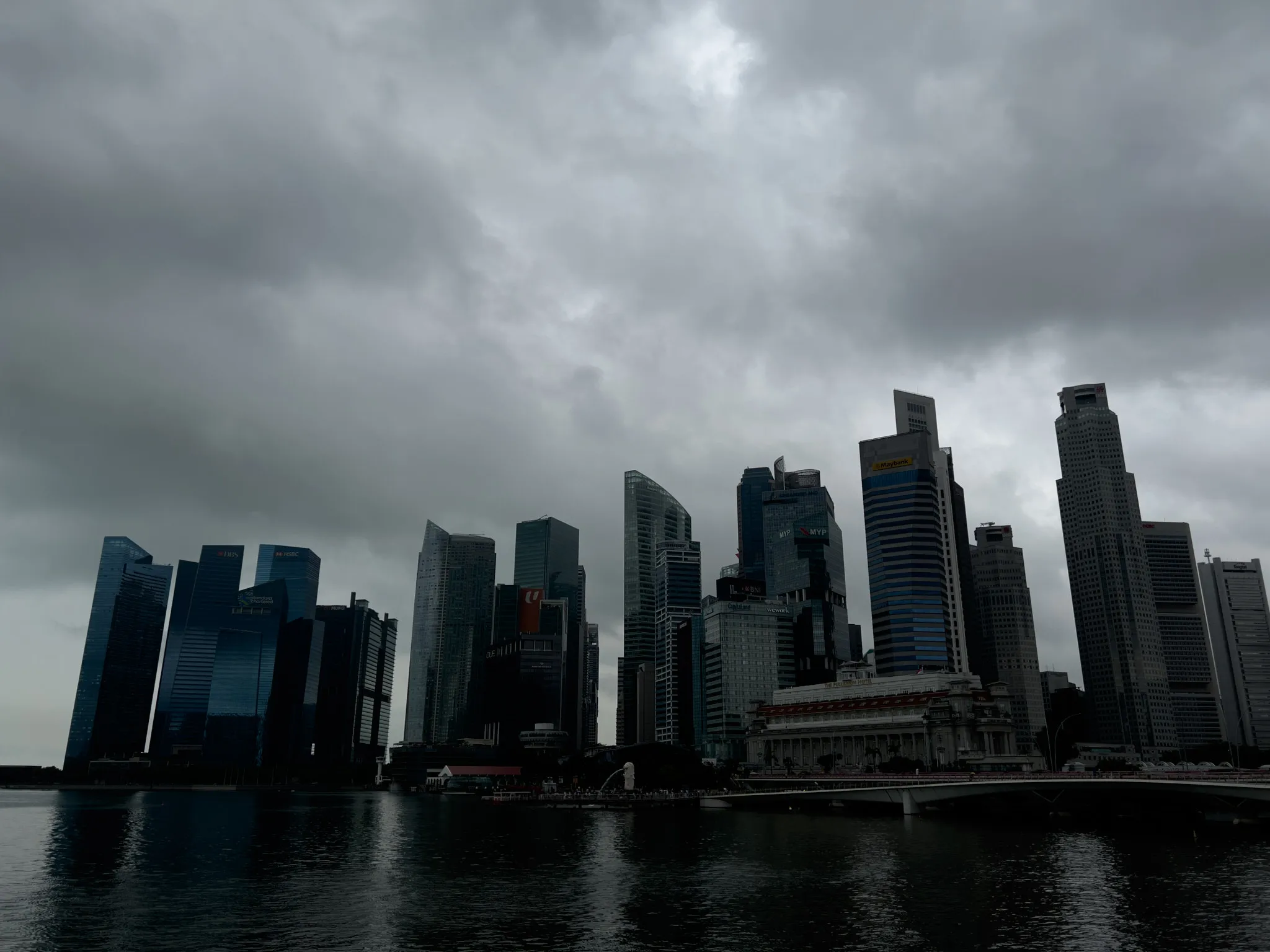 Dense cluster of modern high‑rise buildings rising along a waterfront under dark, heavy clouds, with muted reflections of the structures visible on the calm water below.