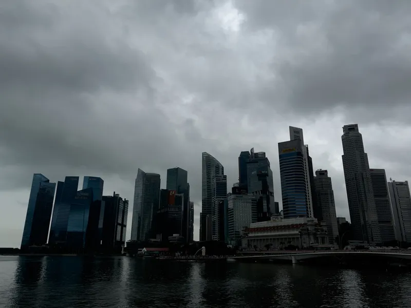 Dense cluster of modern high‑rise buildings rising along a waterfront under dark, heavy clouds, with muted reflections of the structures visible on the calm water below.