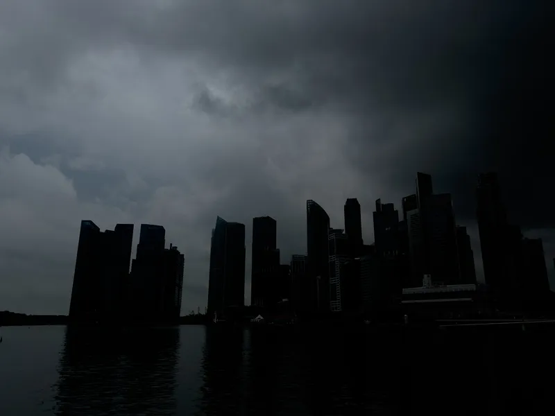 Silhouetted skyscrapers line a waterfront beneath a sky filled with dark, heavy storm clouds. The tall buildings appear nearly black against the dim light, while the water in the foreground reflects the deep shadows and muted tones from above. The overall atmosphere is tense and dramatic, suggesting an approaching storm.