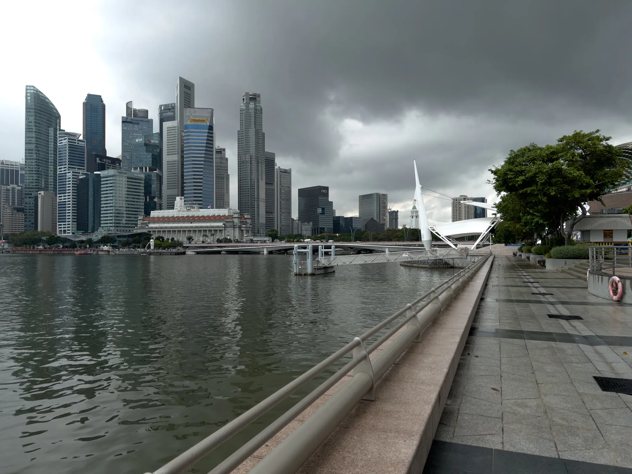 A waterfront promenade stretches along calm, rippling water, leading toward a cluster of modern high‑rise buildings in the distance. The skyline is dominated by tall glass and steel towers, while a white, sail‑like structure stands near the water on the right. Dark, heavy clouds hang low overhead, casting a muted light across the scene. The walkway is mostly empty, lined with trees, benches, and railings, creating a quiet and slightly dramatic atmosphere before an approaching storm.