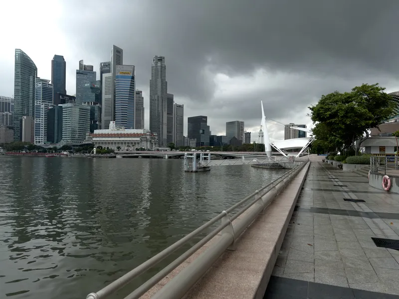 A waterfront promenade stretches alongside calm, rippling water, with a row of modern high-rise buildings forming a dense skyline under heavy, dark storm clouds. A distinctive white architectural structure with angled supports stands near the water’s edge, and a broad stone walkway runs parallel to the water, bordered by trees and railings. The scene has a quiet, atmospheric mood as the incoming weather looms over the cityscape.