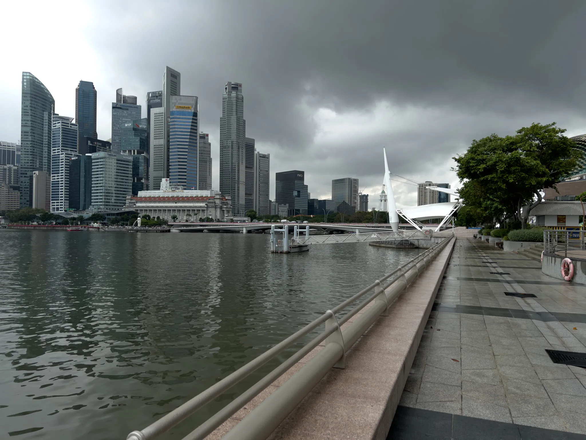 A waterfront promenade stretches alongside calm, rippling water, with a row of modern high-rise buildings forming a dense skyline under heavy, dark storm clouds. A distinctive white architectural structure with angled supports stands near the water’s edge, and a broad stone walkway runs parallel to the water, bordered by trees and railings. The scene has a quiet, atmospheric mood as the incoming weather looms over the cityscape.