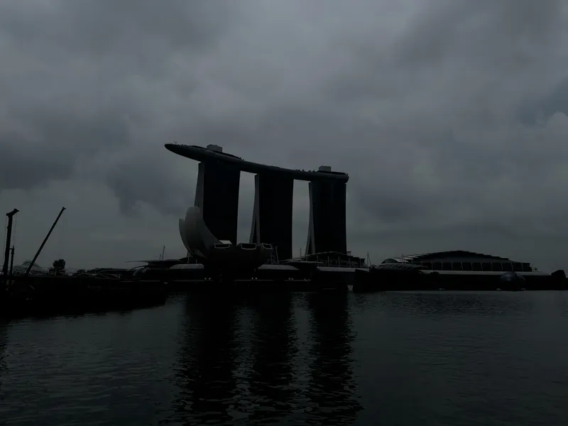 A dramatic waterfront scene dominated by the dark silhouette of a futuristic three‑tower structure connected by a large rooftop platform, set against heavy overcast skies. The calm water in the foreground reflects the shadowed architectural forms, while additional modern buildings and sculptural elements surround the central structure. Thick clouds fill the sky, creating a moody and atmospheric setting.