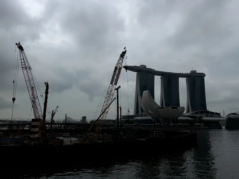 Large construction cranes rise above a waterfront work site under a heavy, overcast sky, with dark clouds diffusing the light across the scene. Behind the cranes stands a distinctive three‑tower structure connected by a sky‑level platform, along with a lotus‑shaped building nearby. The water in the foreground reflects the muted tones of the cloudy atmosphere.