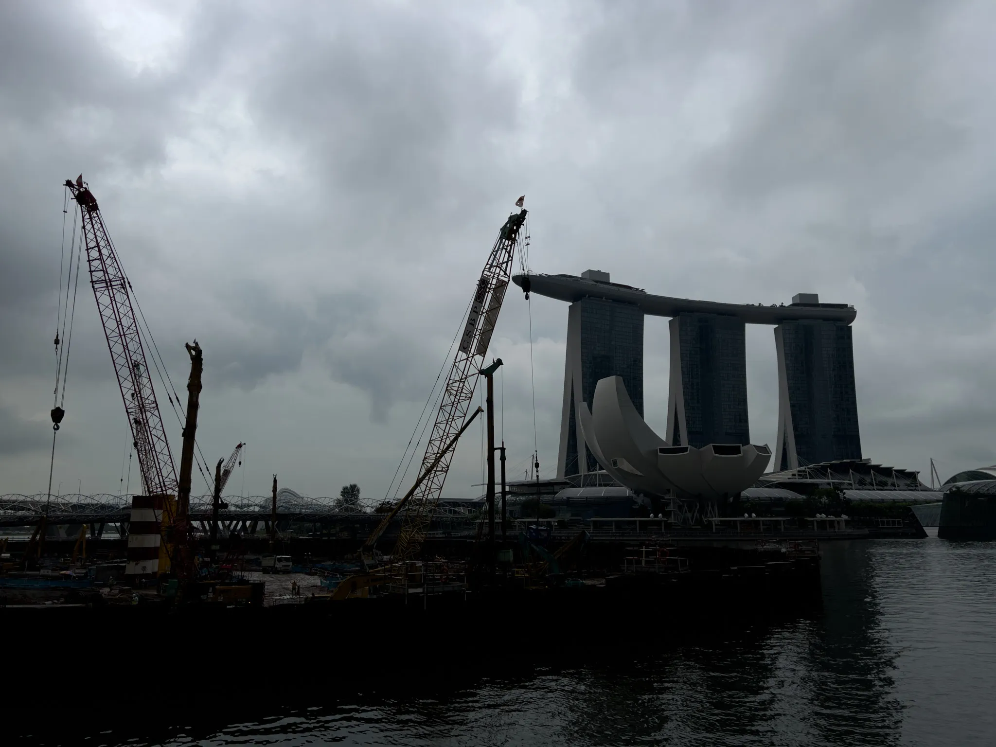 Large construction cranes rise above a waterfront work site under a heavy, overcast sky, with dark clouds diffusing the light across the scene. Behind the cranes stands a distinctive three‑tower structure connected by a sky‑level platform, along with a lotus‑shaped building nearby. The water in the foreground reflects the muted tones of the cloudy atmosphere.