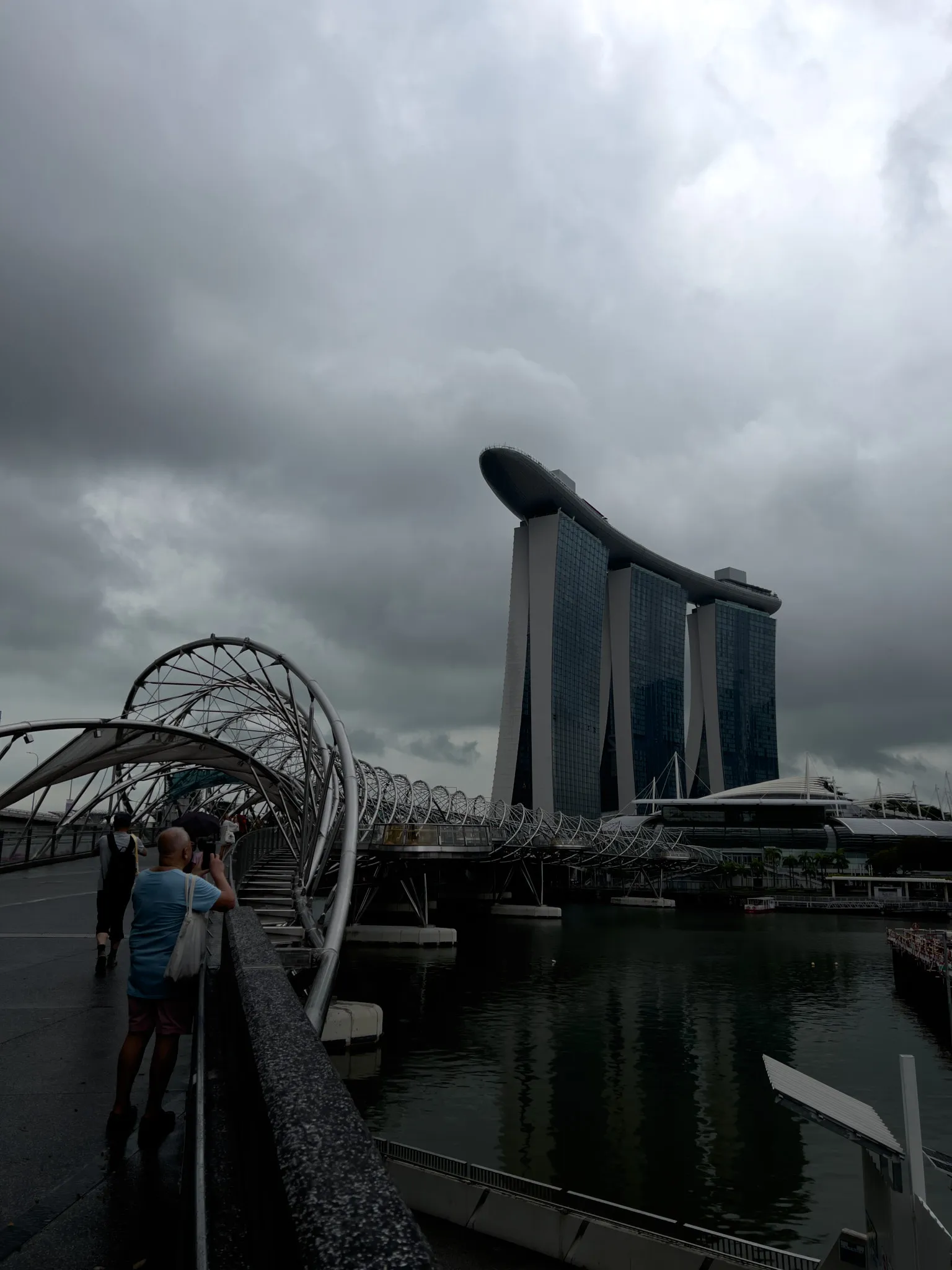 Overcast sky with heavy, dark clouds looming above a waterfront area featuring a modern, three‑tower building topped by a long, curved structure. A metallic pedestrian bridge with a helical design stretches over the water toward the building. People walk along the bridge and the adjacent pathway, including someone taking a photo. The water below reflects the muted light and the architectural forms.