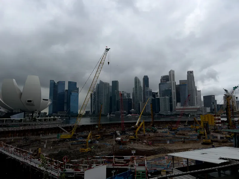 Large construction site filled with cranes, excavators, and heavy machinery set against a dense cluster of modern high‑rise buildings. Dark, heavy clouds hang low over the city, creating a dramatic contrast with the bright yellow construction equipment. A distinctive white, lotus‑shaped building stands to the left, while numerous skyscrapers form the skyline across the water. The ground is busy with workers, scaffolding, and partially excavated areas, suggesting major urban development in progress.