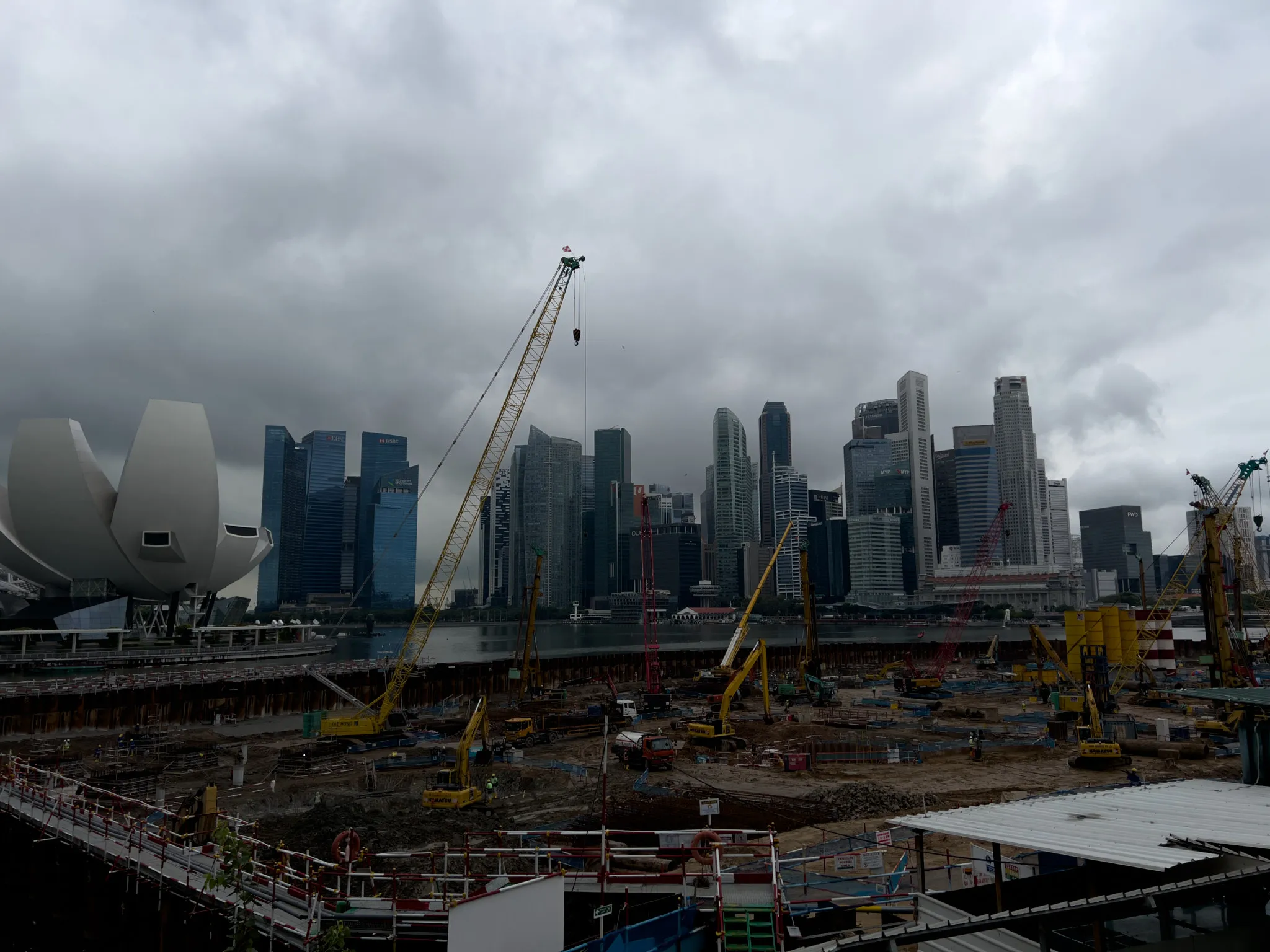Large construction site filled with cranes, excavators, and heavy machinery set against a dense cluster of modern high‑rise buildings. Dark, heavy clouds hang low over the city, creating a dramatic contrast with the bright yellow construction equipment. A distinctive white, lotus‑shaped building stands to the left, while numerous skyscrapers form the skyline across the water. The ground is busy with workers, scaffolding, and partially excavated areas, suggesting major urban development in progress.