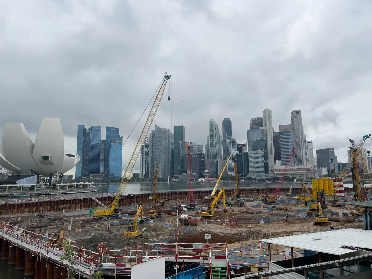In this scene in the Civic District, Downtown Core, Central Region, Singapore, extensive construction is underway. In the foreground, numerous construction machines and cranes are at work on a building site. The site is surrounded by steel beams, while in the background the striking Singapore skyline rises with modern skyscrapers. An overcast sky lends the scene a dynamic and bustling atmosphere. Also visible in the foreground is a distinctive architectural element shaped like a blossom.