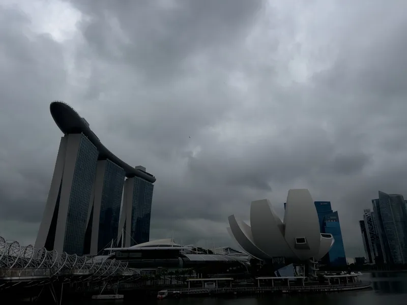 A cluster of futuristic waterfront buildings stands under a sky filled with dark, heavy clouds. A tall structure with three vertical towers connected by a long, curved rooftop platform rises prominently to the left, its glass surfaces reflecting the muted light. To the right, a sculptural, lotus‑shaped building opens outward in smooth, curved petals. Additional high‑rise towers line the background, while a pedestrian bridge with a looping, lattice-like design stretches across the lower left side toward the water.
