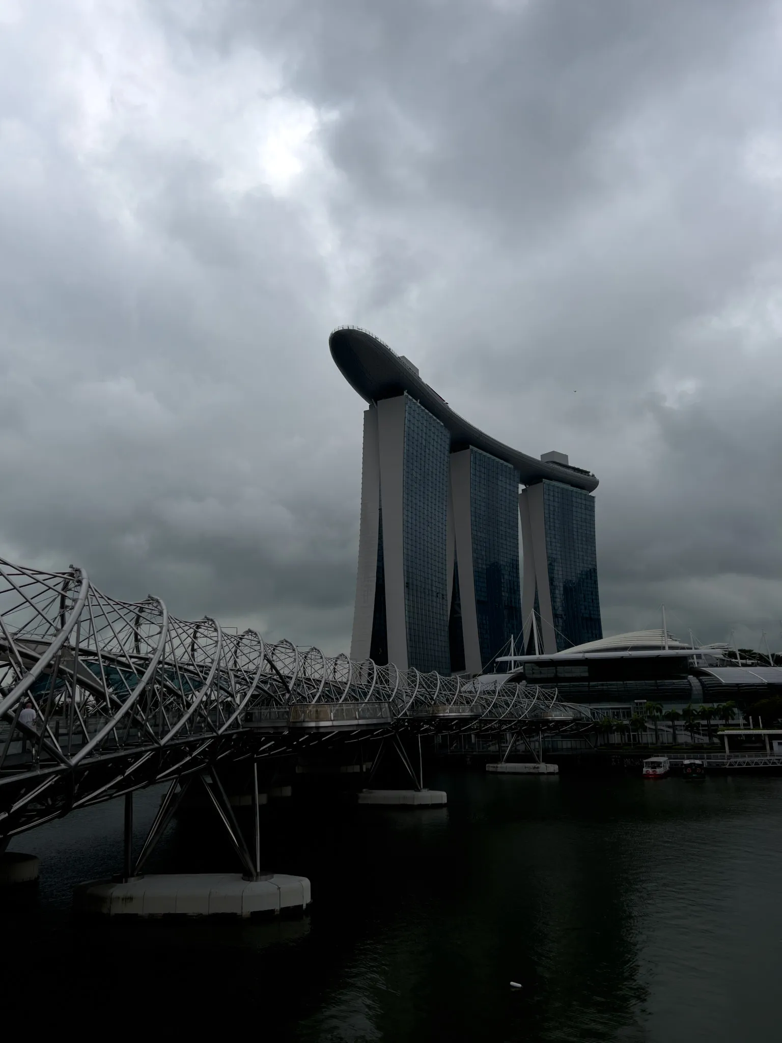 A dramatic, overcast sky looms over a modern waterfront scene featuring a tall, three‑tower structure topped with a long, curved platform. In the foreground, a metallic, spiral‑like pedestrian bridge stretches across the water toward the buildings. Dark clouds fill the sky, casting deep shadows over the water and surrounding architecture.