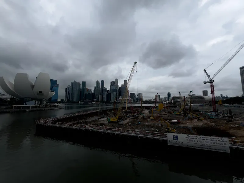 Large waterfront construction site with multiple cranes, machinery, and materials spread across a wide excavation area, set beside calm water. In the background, a dense cluster of modern high‑rise buildings forms a prominent skyline under heavy, dark clouds. A distinctive white, sculptural building stands to the left near the water, while safety signage is positioned along the perimeter of the construction zone.