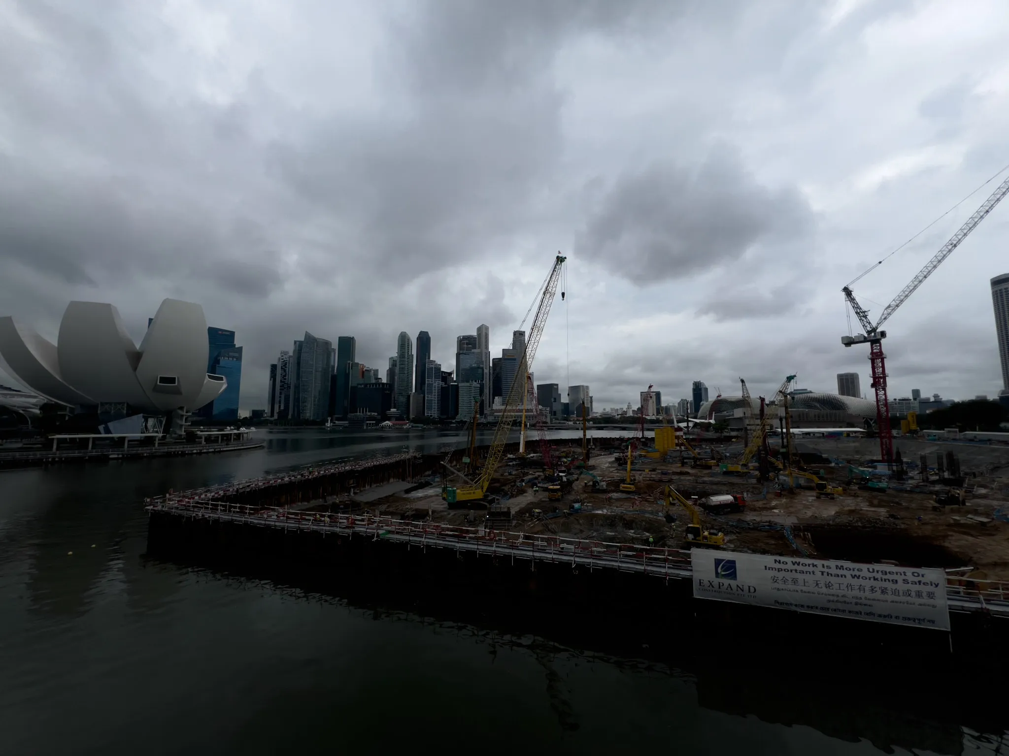 Large waterfront construction site with multiple cranes, machinery, and materials spread across a wide excavation area, set beside calm water. In the background, a dense cluster of modern high‑rise buildings forms a prominent skyline under heavy, dark clouds. A distinctive white, sculptural building stands to the left near the water, while safety signage is positioned along the perimeter of the construction zone.