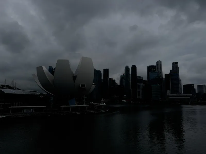A waterfront cityscape under heavy, dark clouds, showing a distinctive lotus‑shaped modern building in the foreground and a cluster of tall skyscrapers forming a dense skyline behind it. The water reflects the darkened tones of the overcast sky, and the overall atmosphere appears moody and stormy.