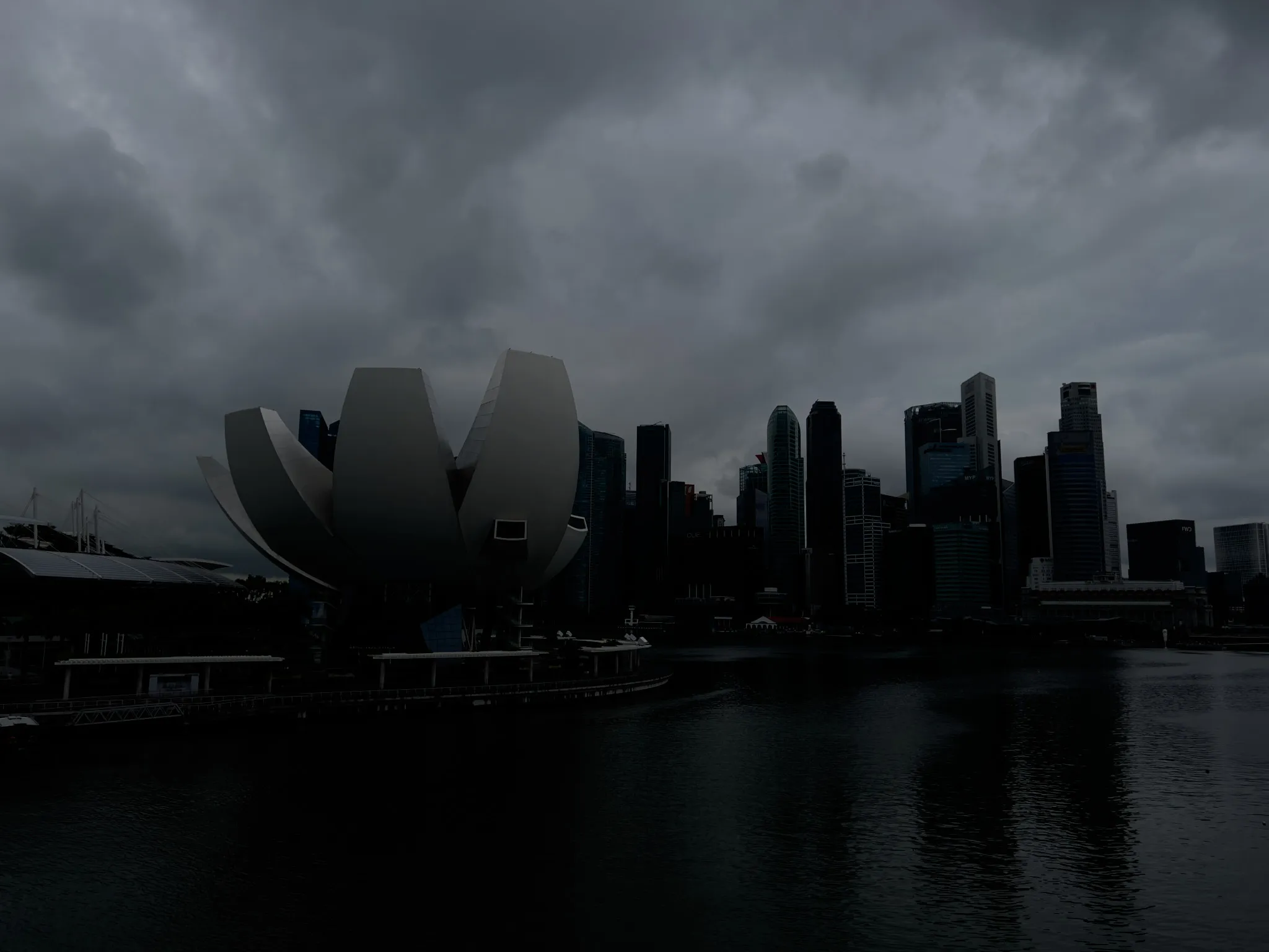 A waterfront cityscape under heavy, dark clouds, showing a distinctive lotus‑shaped modern building in the foreground and a cluster of tall skyscrapers forming a dense skyline behind it. The water reflects the darkened tones of the overcast sky, and the overall atmosphere appears moody and stormy.