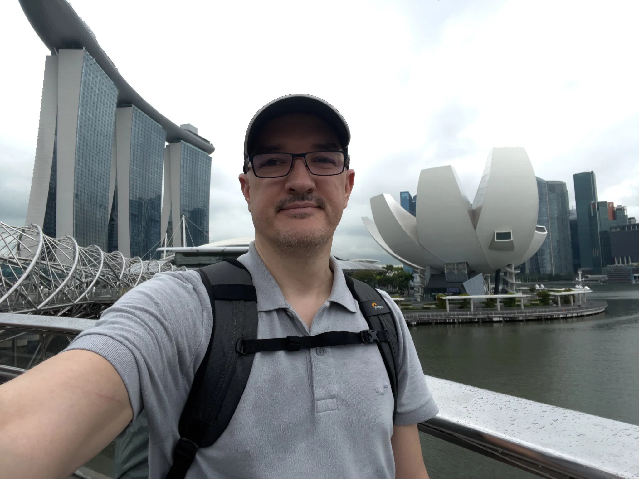 A person taking a selfie on a waterfront bridge with modern architecture in the background, including a three‑tower building topped by a long horizontal structure and a large white lotus‑shaped building near the water. The sky appears overcast, and the bridge railing shows raindrops. The person wears a cap, glasses, a light gray polo shirt, and a backpack.