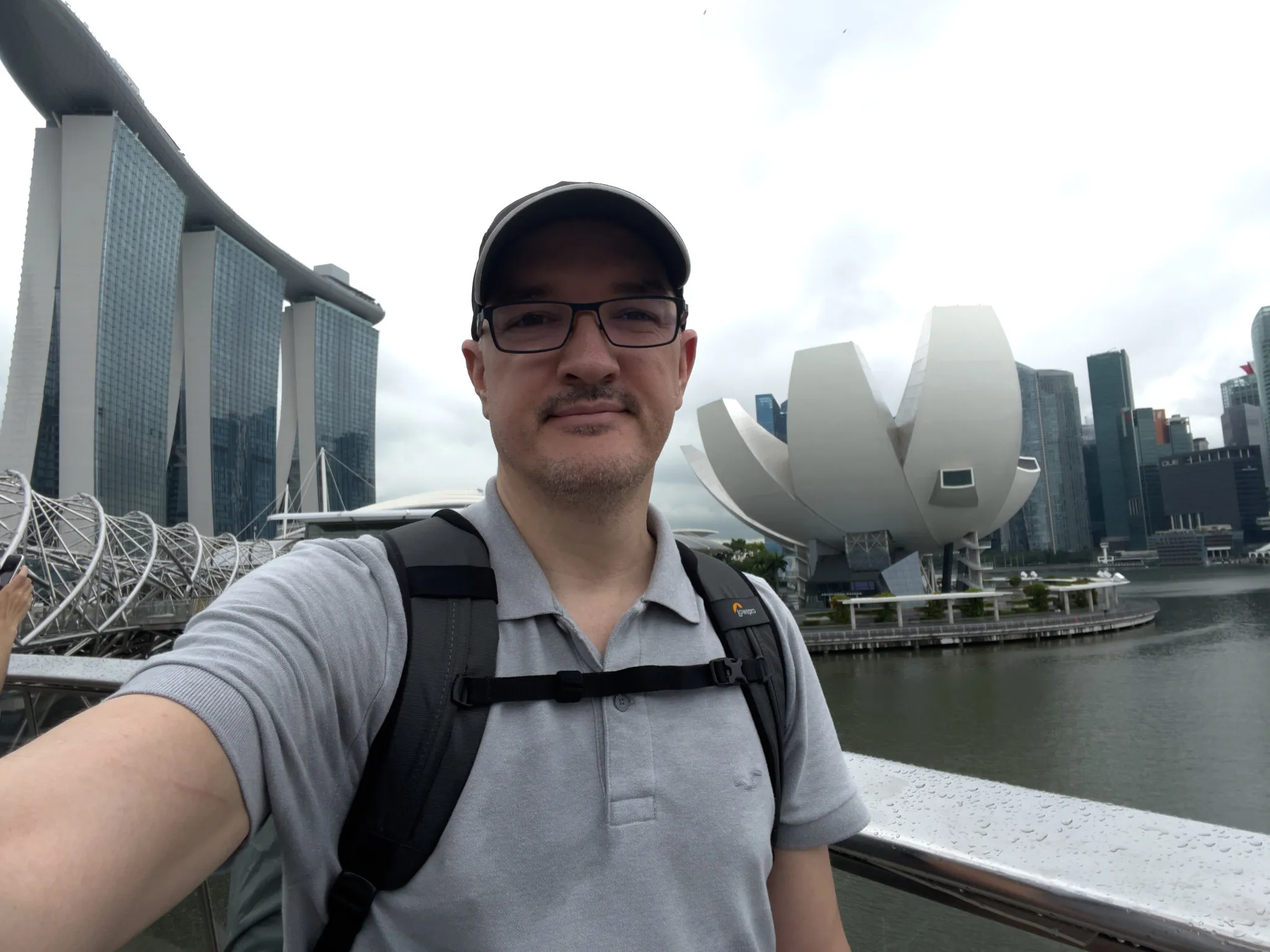 A person wearing a gray polo shirt, black-rimmed glasses, a cap, and a backpack stands on a waterfront bridge with modern architecture in the background. To the left rises a tall three‑tower building topped by a long horizontal structure, and to the right is a white lotus-shaped building set against a cluster of high-rise towers. The sky is overcast, and the water below reflects the surrounding structures.