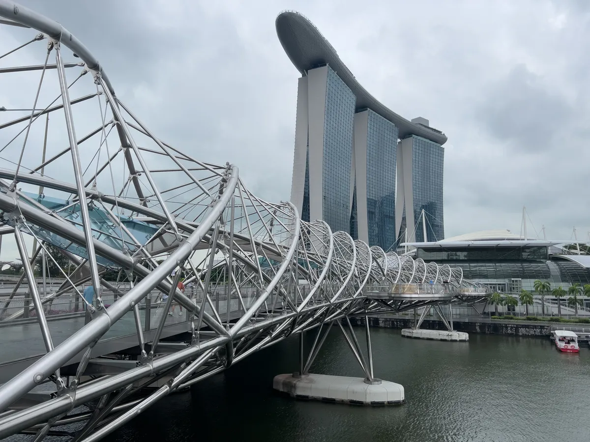 In the Civic District, Downtown Core, Central Region, Singapore, 179803, an elegant, futuristic bridge stretches across the water. Made of gleaming steel, the bridge is defined by its curved, net-like structure that links the two sides of the shore. In the background, the impressive Marina Bay Sands rises, its distinctive architecture and glass facades shaping Singapore's skyline. Grey clouds drift across the sky, lending the scene a dramatic mood.