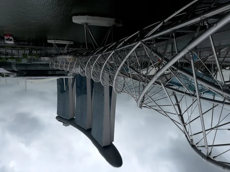 A twisting steel pedestrian bridge with a double-helix design extends over calm waterfront water, leading toward a distinctive three-towered building topped by a long, curved structure. The cloudy sky creates a muted backdrop for the modern architectural elements, and a few people can be seen walking along the bridge.