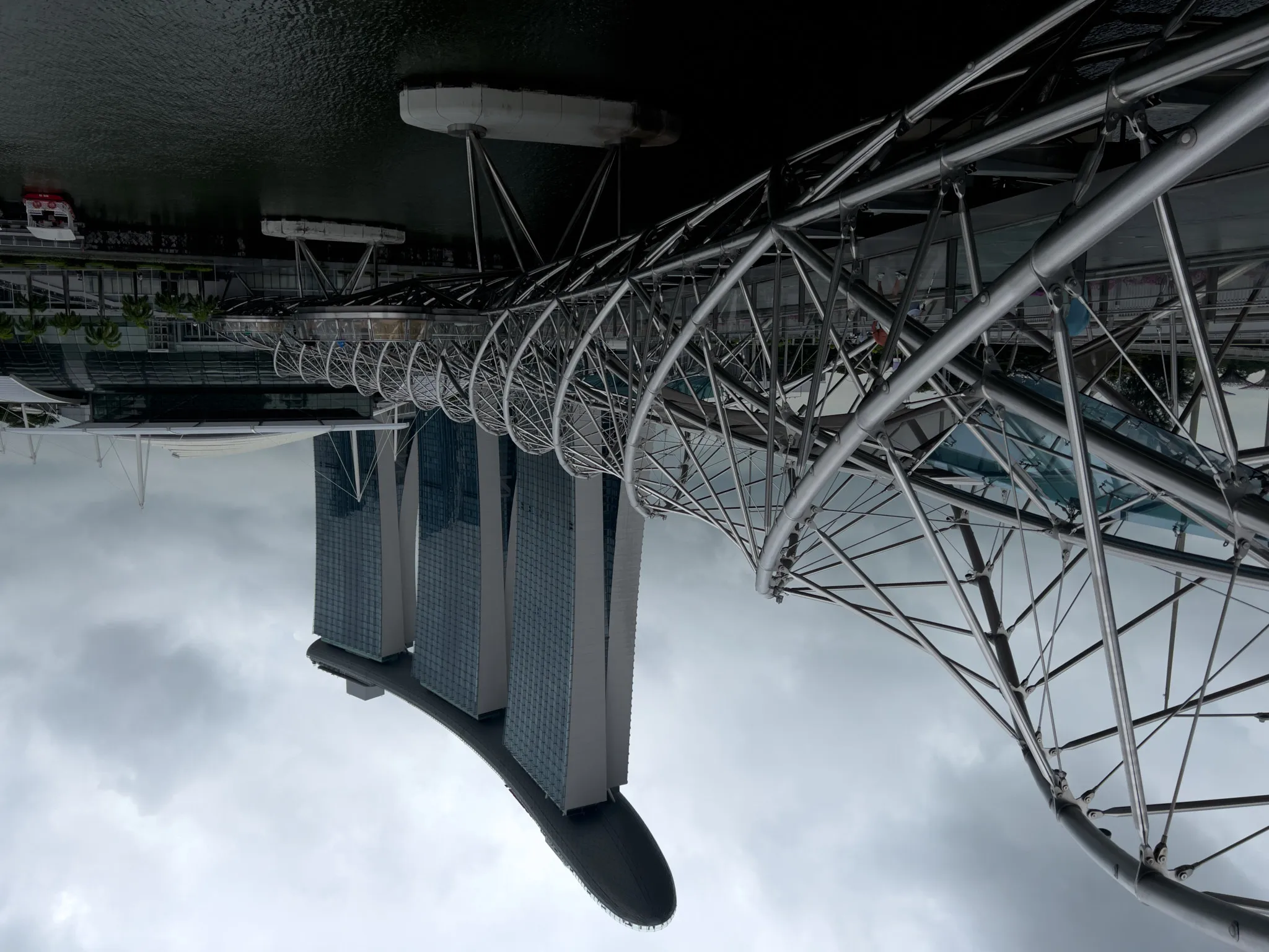 A twisting steel pedestrian bridge with a double-helix design extends over calm waterfront water, leading toward a distinctive three-towered building topped by a long, curved structure. The cloudy sky creates a muted backdrop for the modern architectural elements, and a few people can be seen walking along the bridge.