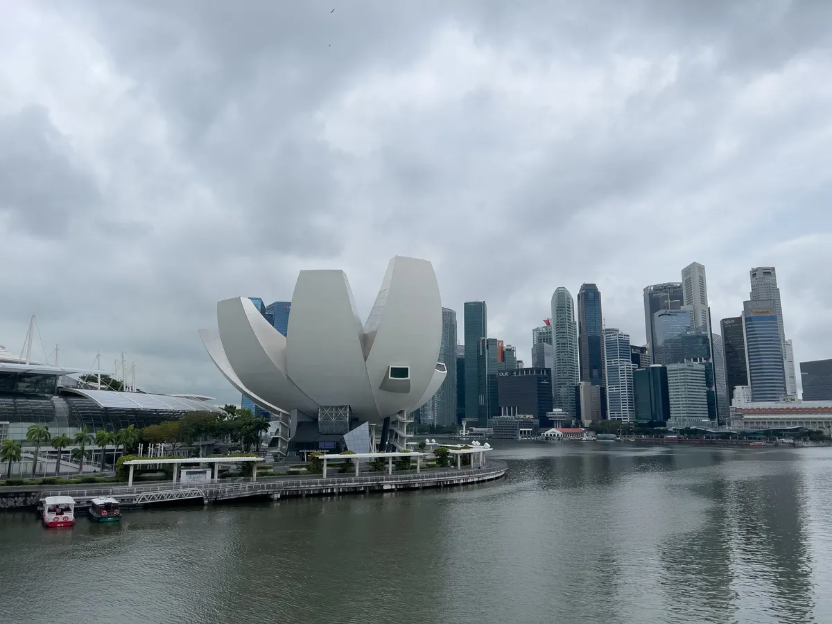 The scene captures the impressive architecture of the ArtScience Museum, shaped like a lotus flower, at the edge of a calm body of water in Singapore. In the background, the modern skyline of the Civic District, Downtown Core, Central Region, Singapore rises up, with tall office buildings reaching above a clear sky behind dense clouds. A few small boats sit on the water, and palms line the promenade, lending the view a tropical atmosphere.