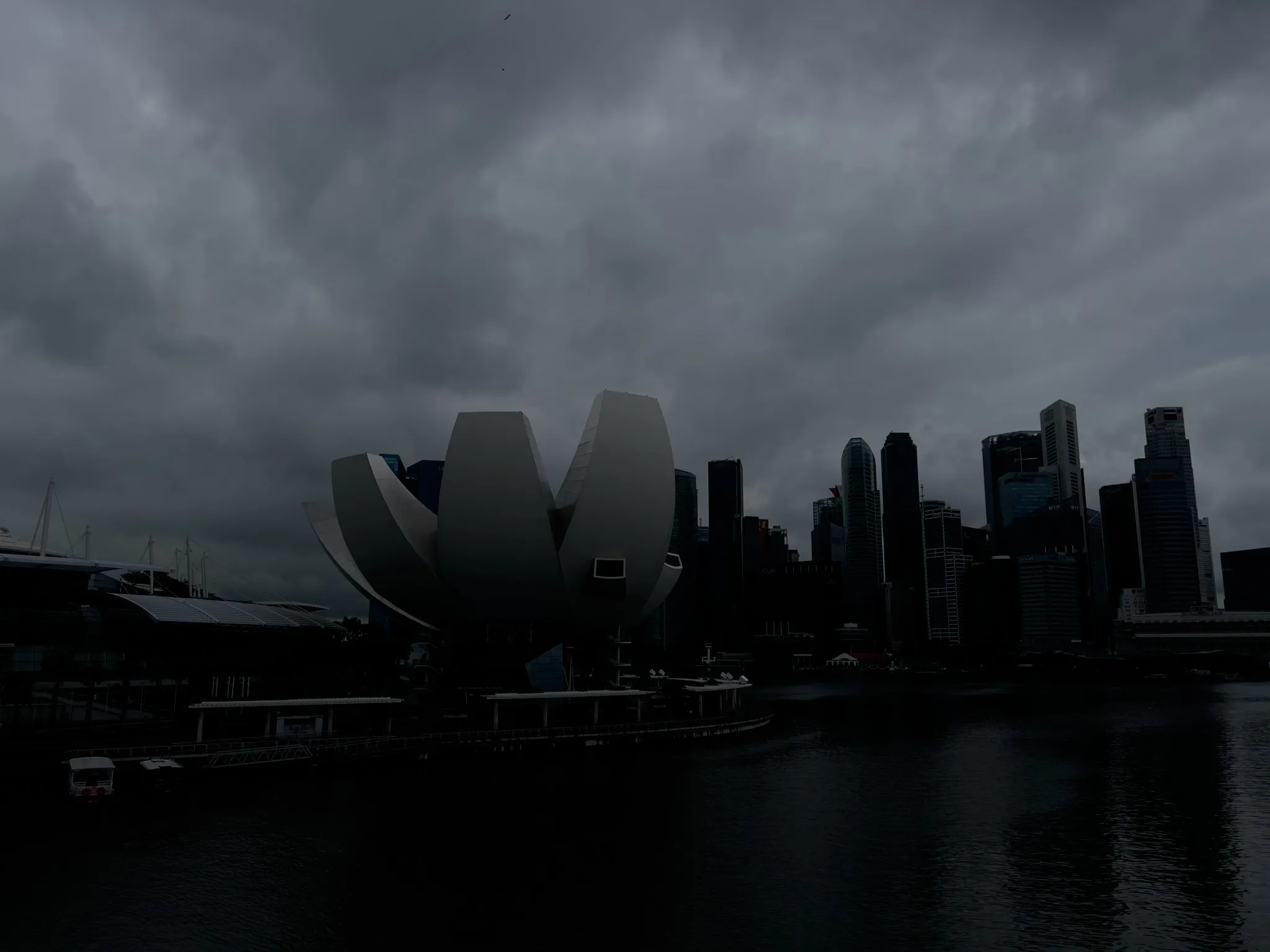 Dark storm clouds gather above a waterfront cityscape, casting deep shadows over the scene. A large, modern, lotus-shaped building stands prominently near the water’s edge, with its curved metallic petals facing the darkened sky. Tall skyscrapers form a dense skyline to the right, their silhouettes contrasting against the heavy clouds. The water below reflects the dim light of the overcast atmosphere, creating a moody and dramatic setting.