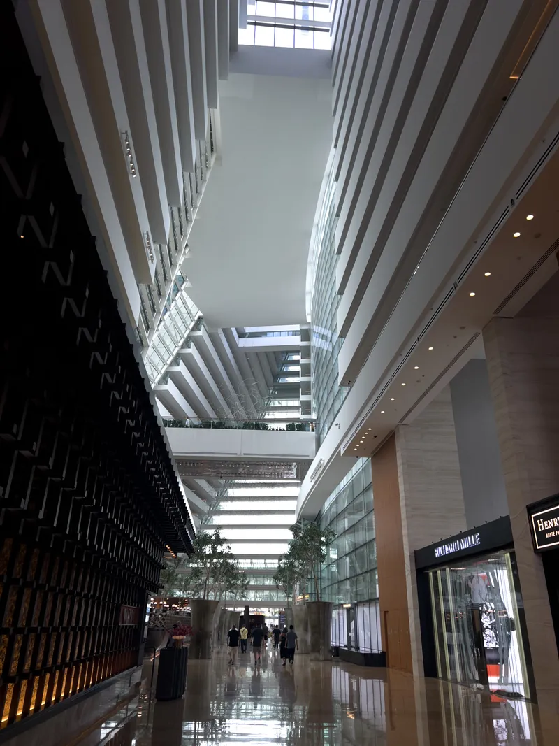 Tall, spacious atrium with tiered balconies rising on both sides, large glass walls allowing natural light to flood the interior, and a glossy floor reflecting the ceiling and surrounding structures. Several people walk along the central corridor lined with shops and trees, while geometric architectural elements create a dramatic sense of height and depth.